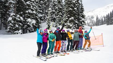 Group of skiers smiles and throws up their poles in celebration on a snowy afternoon with evergreen trees in the background.