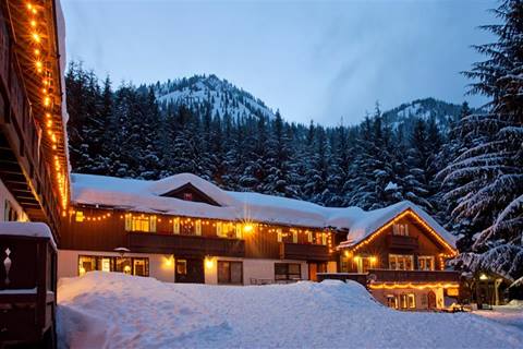Warmly lit, snow-covered mountain lodge with two mountain peaks in the background at twilight. 