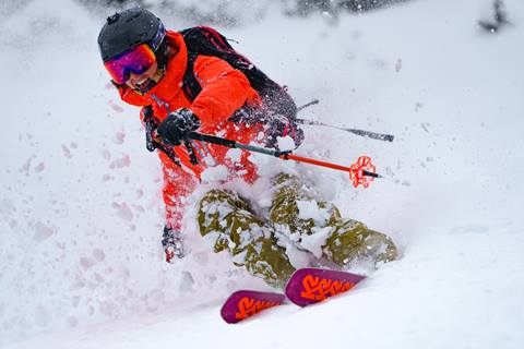 Woman barrels through untouched powder with an orange jacket, beige snow pants, and K2 skis. 