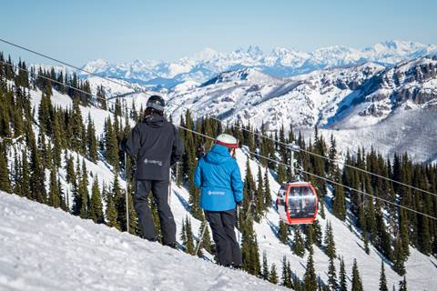Two skiers stop for a break on a mountain side with mountain peaks and a gondola car in the background.