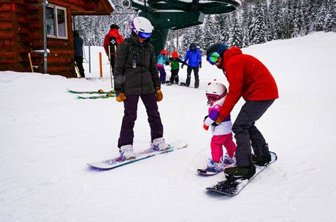 Adult helping a young child practice snowboarding near a chairlift